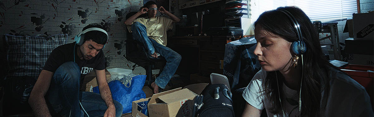 Three people in a dimly lit, cluttered music studio, with two wearing blue AIAIAI Tracks Oklou Edition on-ear headphones.